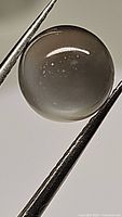 Close-up photo of a round cabochon cut Silver Moonstone gemstone held by tweezers. The stone is translucent with small internal inclusions visible and a glossy surface.