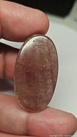 Close-up of oval polished natural strawberry quartz gemstone held between fingers, showcasing reddish-pink color and internal natural inclusions.