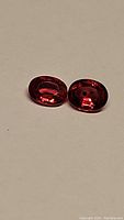 Two oval deep electric red rubies, one with inclusions, displayed on neutral surface under direct lighting to show color and clarity.