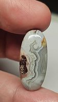 Close-up photo showing oval white crazy lace agate cabochon with banded patterns and druzy section, held between fingers.