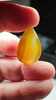 Natural yellow striped agate gemstone held between fingers, backlit to highlight translucent yellow and white banding details in a pear-shaped cabochon cut.