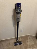 Dyson stick vacuum cleaner standing upright against beige wall and carpet. Clear dust container visible. Power cord included and visible on floor.