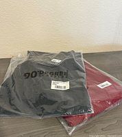 Photo showing two shirts in sealed plastic packages, one black and one red, on a wooden surface.