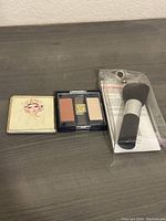 Lot of unopened makeup brush in packaging, and two makeup compacts placed on a gray surface.