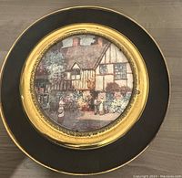 Front view of vintage brass wall hanging plate showing central cottage scene with two women and floral garden surroundings, set on black border with brass rim.