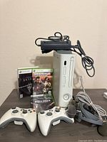 Front view of white Xbox 360 console with power supply placed on top. Two white wireless controllers in front. Three Xbox 360 game cases visible behind: Skyrim, Mass Effect 2, Call of Duty Black Ops. Wired headset with microphone also present.