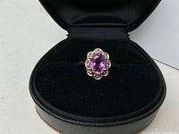Front view of ring in case showing oval purplish-pink center stone and halo of white accent stones