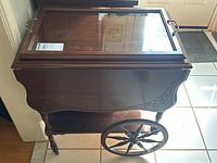 Front view of walnut tea cart showing hinged glass top, brass handles and carved apron.
