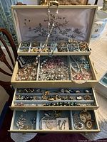Opened jewelry box showing top compartment with small cubbies holding brooches and earrings, strands of beads draped