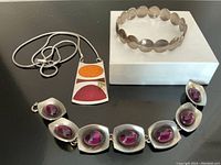 Three jewelry pieces on black surface: sterling silver orb bangle on white box, Jensen pewter bracelet with purple stones, Silvestri de Passile enamel pendant necklace.