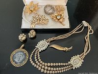 Overview on black surface showing multi-strand necklace, monogram pendant, cluster earrings, curved leaf brooch and five brooches laid in box