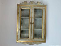 Front view of gold and gray-blue painted olive wood wall cabinet with mesh doors and scrolled crest