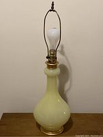 Full view of a vintage table lamp with yellow glass bulbous body, brass base, brass fittings, white light bulb, and harp for lampshade attachment.