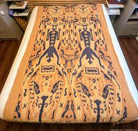 Full view of large decorative ikat textile blanket with two large warrior figures and accompanying mythological and natural symbols in indigo on beige background.