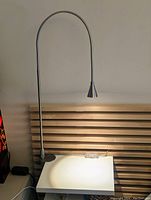 Silver metal adjustable neck lamp clamped to a white surface against a wooden slatted background, lit and casting light downward.