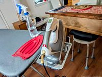 Black & Decker iron standing on a metal ironing board with gray cover. A red silicone iron rest and spray water bottle are placed on the board.