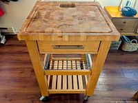 Front view of the kitchen island showing the butcher block top with groove and central cutout, drawer with handle, metal wire rack, and slatted lower shelf.