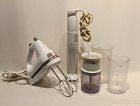 Photo shows two small kitchen hand mixers and accessories on a plain background. KitchenAid 5-speed mixer with beaters on left, Braun hand mixer with chopper attachment and two plastic measuring containers on right.