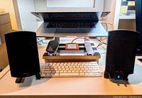 Two small black desktop Klipsch speakers positioned on either side of a white keyboard with a laptop on a stand above them.