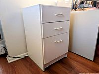 Side angle photo of the white 3-drawer cabinet showing its height and depth, placed on wood floor.