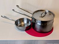 Two stainless steel cooking pots side by side on a white surface with a red circular mat underneath. Larger pot has lid and steamer basket inside, smaller pot is a sauce pot.