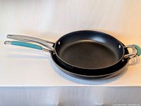 Two non-stick frying pans stacked; 12-inch Calphalon on top, 14-inch Rachael Ray below, showing their size difference and handle design.