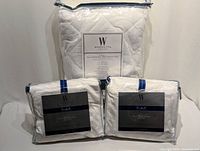 Full view of two Wamsutta sheet sets and one waterproof mattress pad in original unopened packaging, white color.