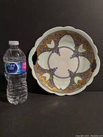 Photo showing the front of a round serving plate with an Art Nouveau grape and gold leaf motif and scalloped edge next to a water bottle for size reference.