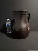Front and angled view of vintage glazed stoneware jug next to a water bottle for size reference. Jug is dark brown with a glossy finish and has a handle and wide mouth.