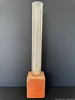 Single photo of a large brutalist abstract sculpture with a clear cylindrical tube mounted on a rusty metal frame and a square brick base against a dark background.