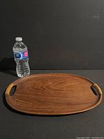 Oval wood platter alongside a standard plastic water bottle to show size and scale. The platter is medium brown with visible wood grain and integrated cut-out handles on each end.
