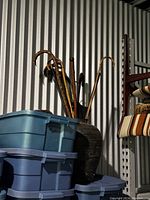 Photo showing seven vintage canes and walking sticks placed upright inside a large ceramic vase, against a corrugated metal wall backdrop with some stacked plastic bins visible to the left.