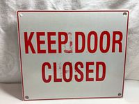 Side of the sign with red text 'KEEP DOOR CLOSED' on white background, with mounting holes in each corner and some signs of wear.