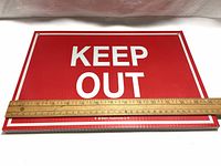 Single red rectangular "KEEP OUT" sign laid flat with wooden ruler showing width about 14 inches.