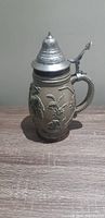 Closed view of West German beer stein showing relief figures, ceramic body, and pewter lid with thumb lift lever.