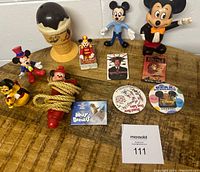 Lot of Disney collectibles displayed on wooden surface: six Disney character figurines, an ice cream cone shaped cup, coiled jump rope with red handles, and four vintage Disney buttons.