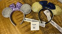 Photo of two Disney Parks 50th Anniversary ears headbands, showing silver glitter ears with a purple bow and gold glitter ears with a dark blue bow, with tags attached.