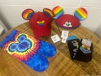 Photo showing the full Disney Pride bundle including the red hat with rainbow ears, the separate rainbow Mickey Mouse ears headband, and tie-dye Mickey Mouse-themed socks