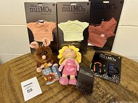 Photo showing three Disney nuiMOs outfits packaged on backing cards in peach, green and red colors, two plush toys, and a Disney Wishables pack arranged on a wooden floor.