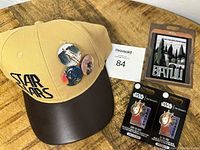 Tan and brown Star Wars hat with four Star Wars buttons pinned on, Batlu patch, and two Star Wars pins in packaging on wooden surface.