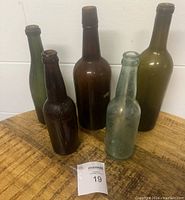 Five vintage colored glass bottles of various heights and colors (brown, green, clear) arranged on a wooden surface with a tag number 19 visible.