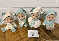 Full view of 4 vintage Christmas ceramic figurines featuring children in holiday outfits holding ornaments and a snowball, arranged in one row on a beige surface in front of a light wall.