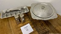 Four vintage kitchen items displayed on a wooden surface: metal ice tray, tin covered dish with floral designs, and salt and pepper shakers.