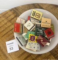 Top view of white milk glass Pyrex pie plate filled with assorted vintage matchbooks showing various advertising designs.