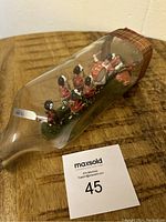 Angled view of the glass bottle showing the Scottish bagpipe band figurines inside, some fallen figurines visible, with a tartan ribbon on the bottle neck.