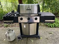 Front view of Broil King propane BBQ grill with two propane tanks and BBQ tools on side shelf.