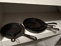 Five cast iron frying pans stacked on a white shelf showing their sizes and handles