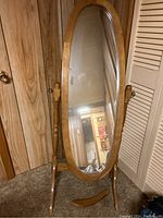 Full view of the cheval mirror showing oval glass and wooden frame, mounted on wooden supports with one horizontal base piece detached and on carpet.