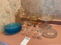 Photo showing the complete lot on a pink surface with six blue glass plates stacked, clear glass compote with lid, two clear glass canisters with lids, and eight stemware glasses arranged in front. Nicotine staining visible.