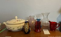 Photo showing the Royal Doulton serving dish with lid, ruby red glass creamers, vintage pink glass pitcher, blue satin glass vase, and clear crackled glass vase arranged together on a wooden surface.